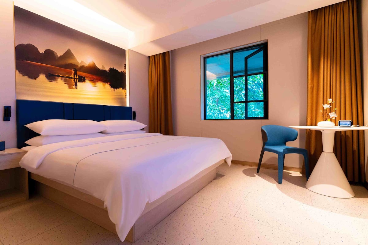 Une chambre d’hôtel comprenant un lit, un bureau, une chaise, une fenêtre donnant sur un espace verdoyant et une fresque murale.