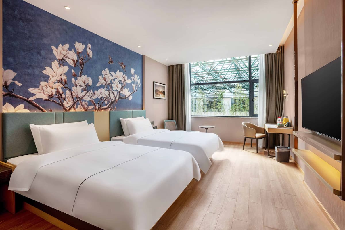 Une chambre d’hôtel avec deux lits, un bureau, une chaise, une télévision et une grande fenêtre donnant sur un paysage verdoyant.