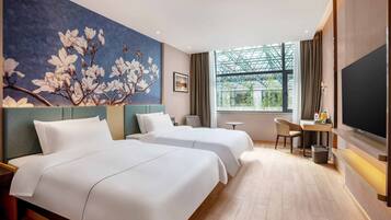 A hotel room with two beds, a desk, a chair, a TV, and a large window with a view of greenery.