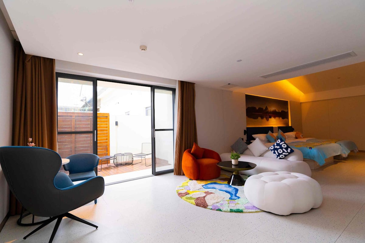 Une chambre d’hôtel moderne dotée d’une grande porte-fenêtre coulissante donnant sur un espace extérieur, d’un ensemble de canapés, d’un tapis rond et d’un lit agrémenté d’un tableau représentant un paysage urbain.