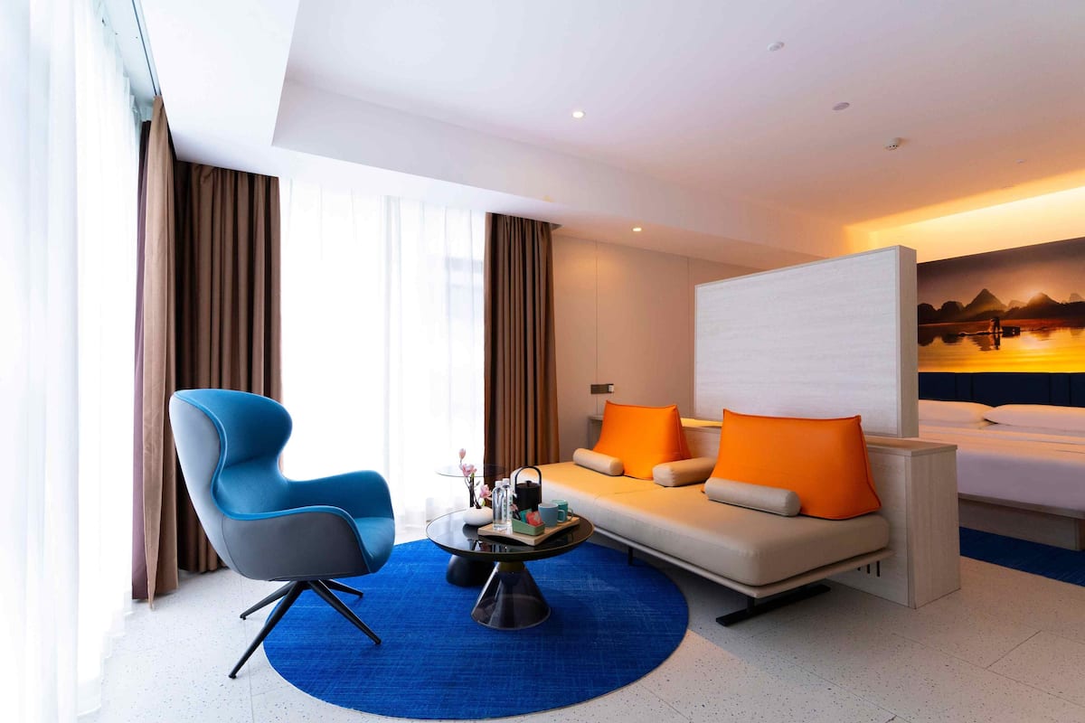 Une chambre d’hôtel moderne avec un canapé, un fauteuil bleu, une table basse et un lit agrémenté d’oreillers orange.