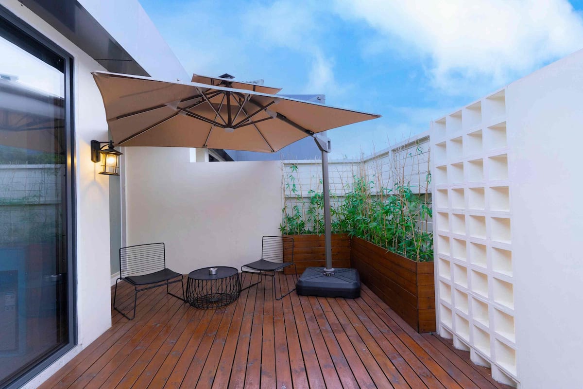 Une terrasse en bois avec une table et des chaises, un grand parasol et un mur orné d’un motif en forme de grille.