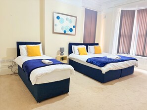 Ein Hotelzimmer mit zwei Betten, jeweils mit blauem Kopfteil, weißer Bettwäsche und gelben Kissen. An der Wand hängt ein Gemälde und ein Fenster mit Vorhängen.