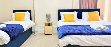 Une chambre d’hôtel avec deux lits, chacun doté d’une tête de lit bleue, de draps blancs et d’oreillers jaunes. Une petite table de chevet en bois, sur laquelle se trouve une lampe et une plante en pot, est placée entre les deux lits.