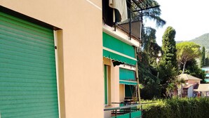 A multi-story building with balconies, a green awning, and a street view with parked cars and greenery.