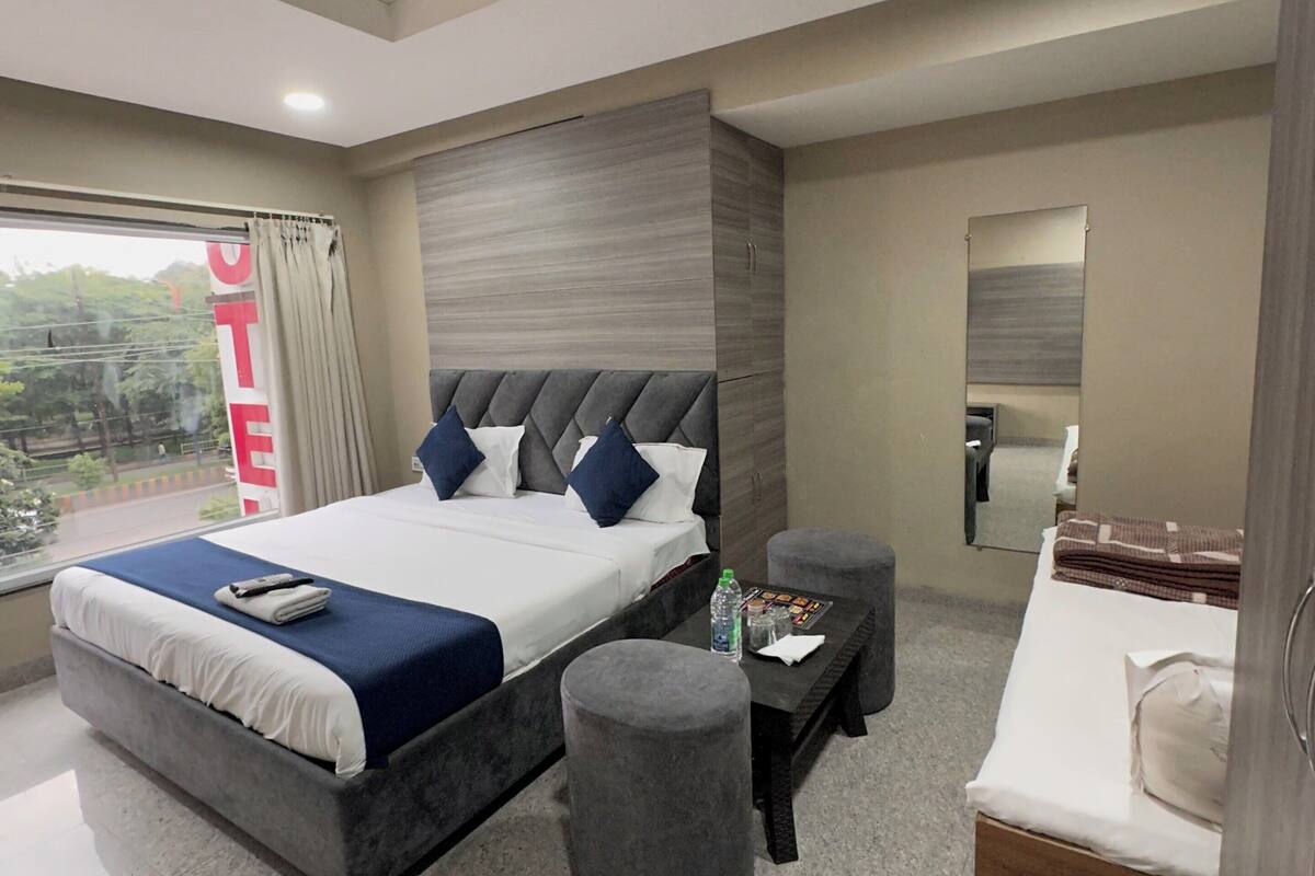 A modern hotel room with a large bed, a desk, a chair, a small ottoman, a ceiling fan, and a large window with a view.
