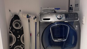 A compact laundry area with a front-loading washing machine, a mop, a broom, and a large bottle of cleaning solution.