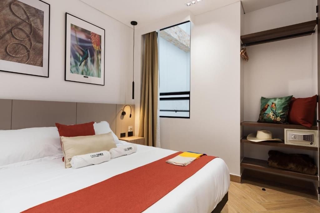 Una habitación de hotel con una cama, una mesita de noche, una lámpara, un estante con un sombrero y una ventana con vistas.