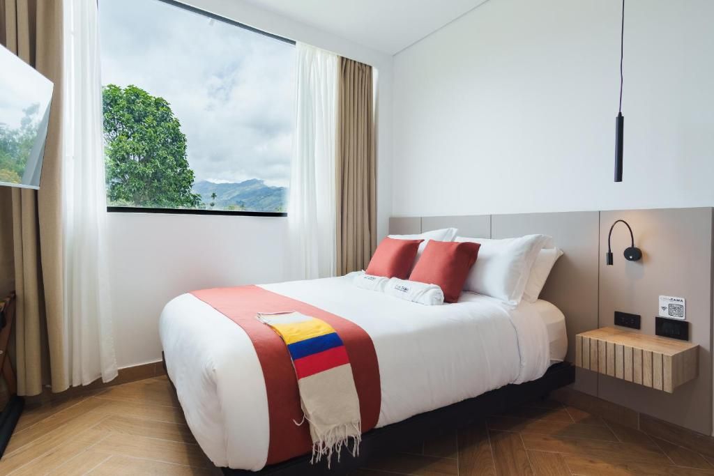 A hotel room with a bed, a window with a view of greenery, a bedside table with a lamp, and a wall-mounted light fixture.