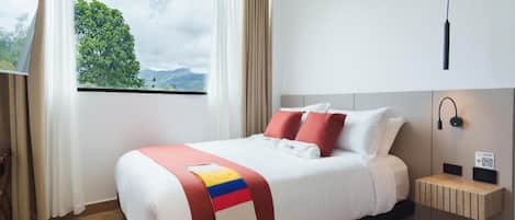 A hotel room with a bed, a window with a view of greenery, a bedside table with a lamp, and a wall-mounted light fixture.