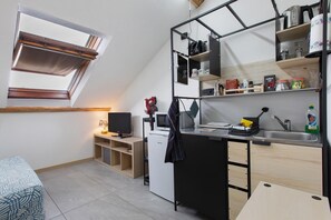 A compact kitchen with a built-in microwave, a sink, and a small refrigerator. There is a window with blinds, a desk with a computer, and a chair.