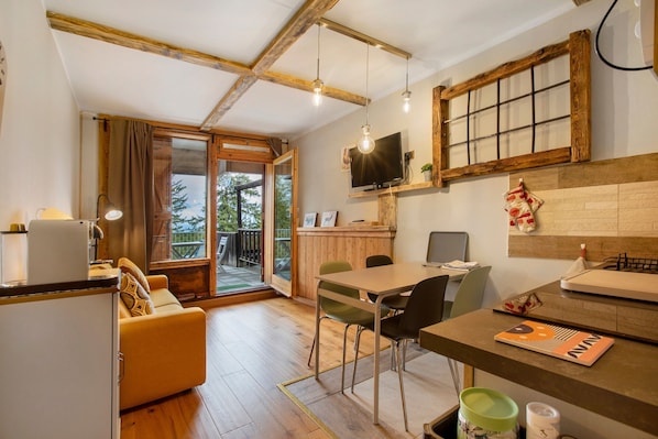A modern kitchen with wooden beams, a dining area with a table and chairs, a sofa, and a TV mounted on the wall.