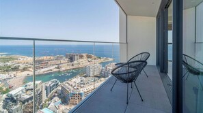 Un balcón con dos sillas de alambre negras, vista despejada al mar y al paisaje urbano, y fachada de un edificio moderno.