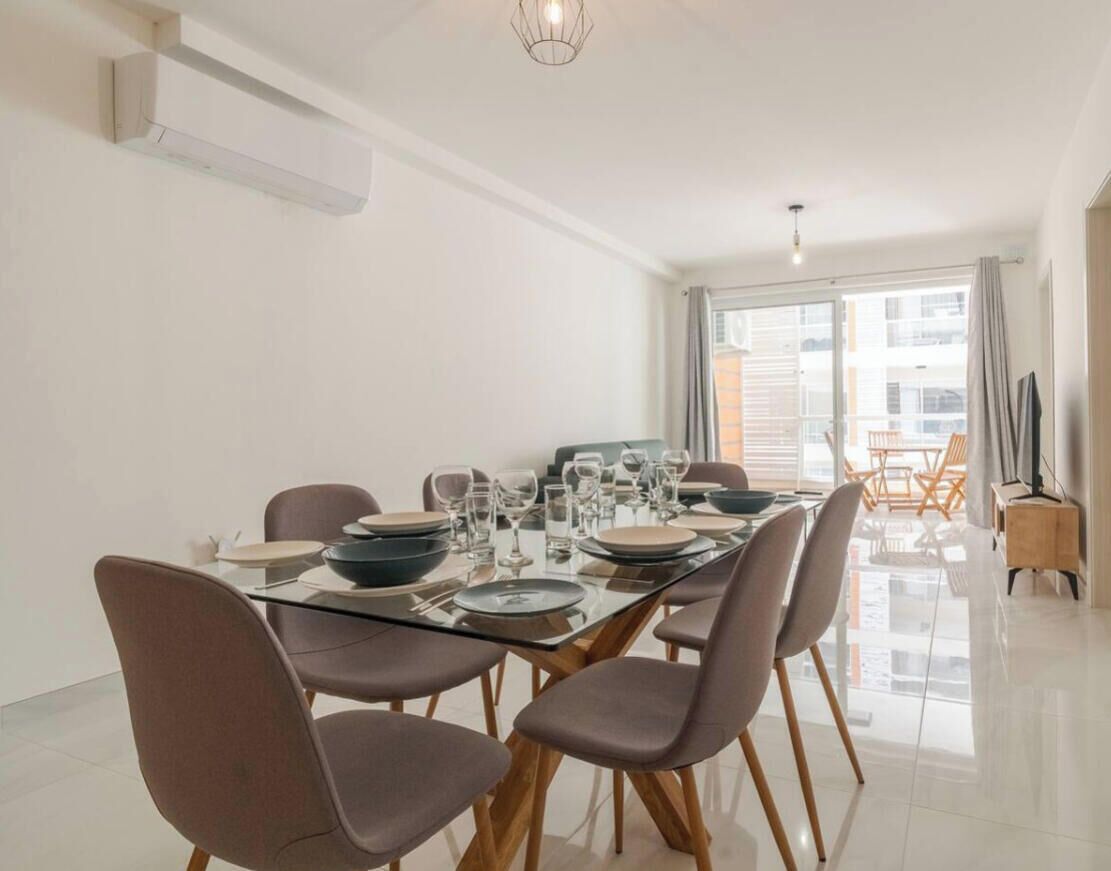 A modern dining area with a long table set for four, surrounded by chairs, in a bright room with large windows and a view of the city.