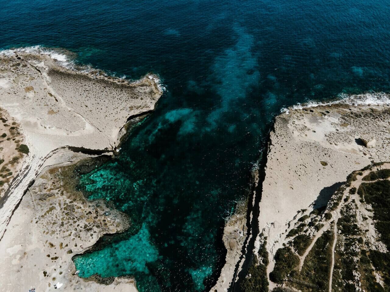 An aerial view of a coastal area with clear turquoise waters, rocky terrain, and sparse vegetation.