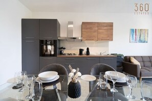 A modern kitchen with grey cabinets, a wooden upper cabinet, and a dining area with a glass table set for six. - Beautiful, fully equipped 2BR home in Mellieha (Mellieħa)