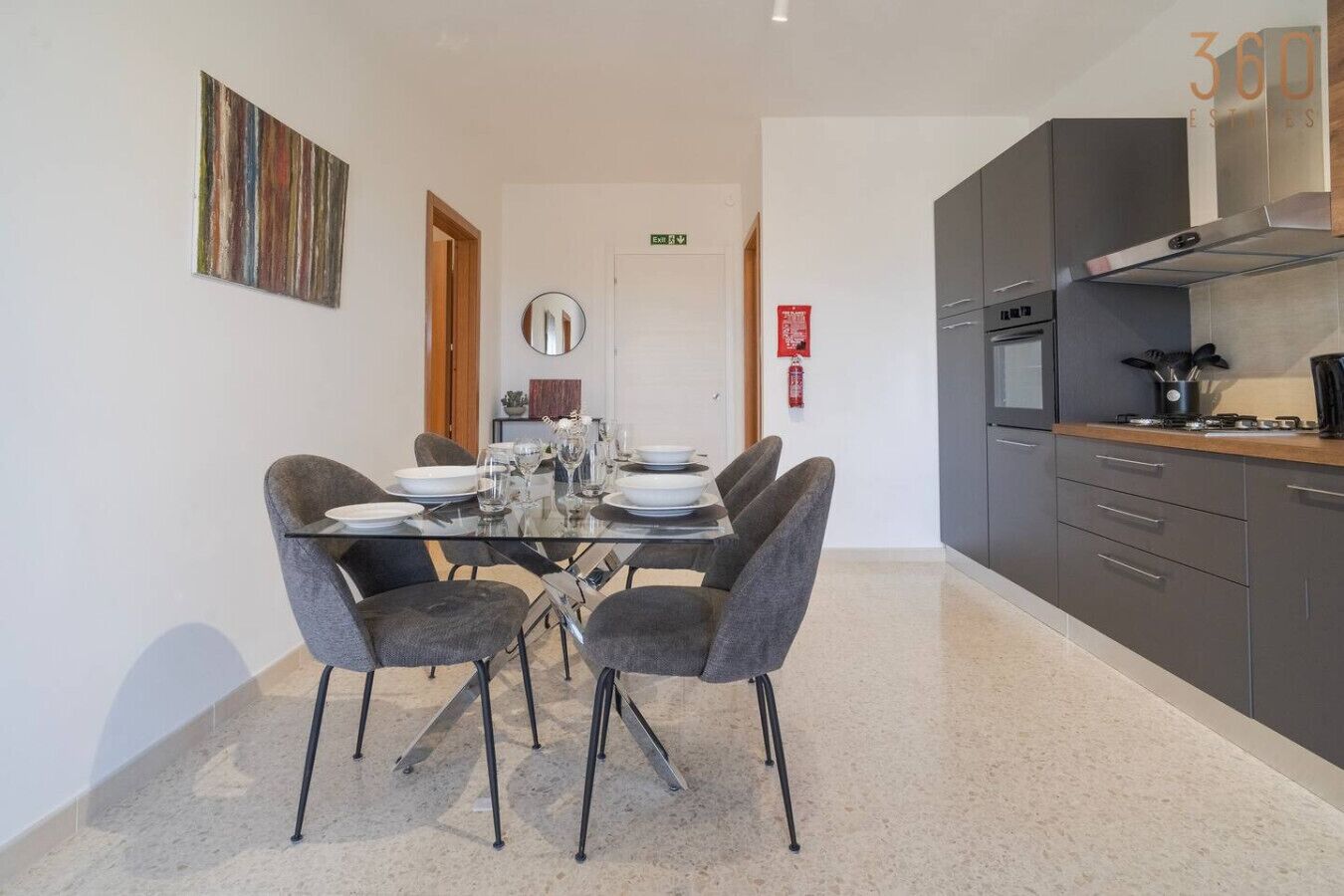 A modern dining area with a glass table and grey chairs, a kitchen with grey cabinets and stainless steel appliances, and a painting on the wall.