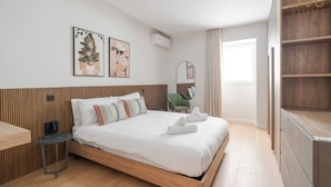 Ein modernes Hotelzimmer mit einem hölzernen Kopfteil, einem Bett mit weißen Bettwäsche, einem kleinen Nachttisch, einem grünen Sessel und einem großen Fenster mit Vorhängen.