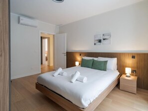 A modern hotel room with a large bed, wooden headboard, bedside lamps, and a view of the hallway.