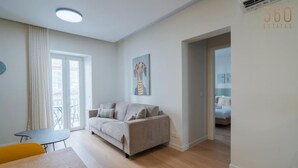 Een moderne woonkamer met een bank, een klein tafeltje en een stoel. Er hangt een schilderij aan de muur en een deur leidt naar een andere kamer.