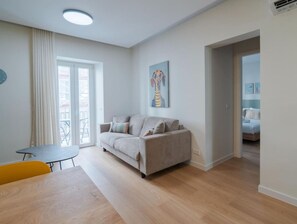 A modern living room with a sofa, a small table, and a chair. There is a painting on the wall and a door leading to another room.
