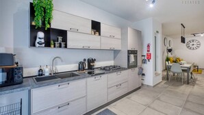 A modern kitchen with grey cabinets, a built-in stove, and a coffee machine. There is a small plant on the wall and a dining area with a yellow table and chairs.