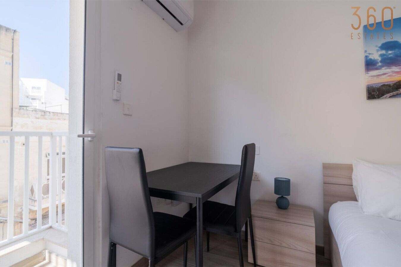 A small room with a table and chairs, a bed, a wall-mounted air conditioner, and a painting.