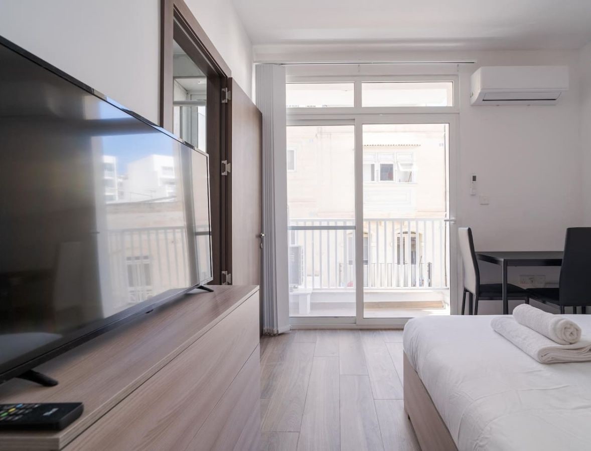 A hotel room with a bed, a desk, a chair, a TV, and a balcony with a view of buildings.