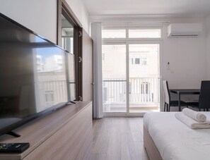 Een hotelkamer met een bed, een bureau, een stoel, een televisie en een balkon met uitzicht op gebouwen.