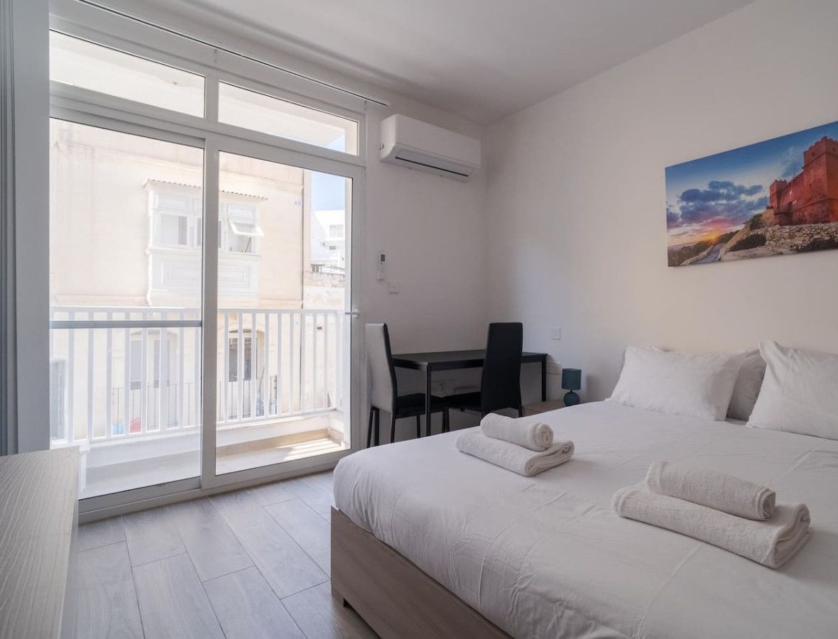 Habitación de hotel con cama, escritorio y silla. Tiene un balcón con vistas a edificios.