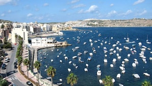 Paisagem costeira com vários barcos na marina, uma estrada notável com palmeiras e edifícios ao longo da orla.