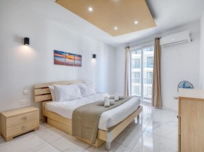 A modern bedroom with a wooden bed, a bedside table, a nightstand, a large window with curtains, and a ceiling light. - Seafront home opposite beach w/WIFI & AC in Qawra (St. Paul`s Bay)