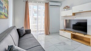 A modern living room with a grey sofa, a flat-screen TV on a wooden stand, and a large window with sheer curtains.