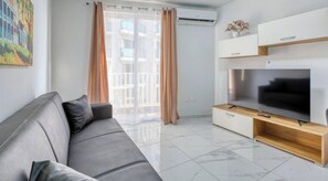 A modern living room with a grey sofa, a flat-screen TV on a wooden stand, and a balcony with curtains.