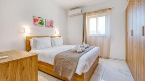 A bedroom with a wooden bed, white bedding, a bedside table, a remote control, a wall-mounted air conditioner, and a window with sheer curtains.