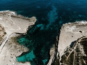 Vista aèria d'una zona costera amb aigües turqueses cristal·lines, terreny rocos i vegetació escassa.