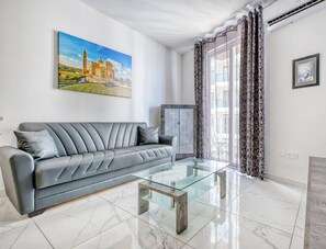 A modern living room with a grey sofa, a glass coffee table, and a painting of a building on the wall. - Sun, Sea & Comfort, Qawra Home w/ Stunning Views (St. Paul`s Bay)