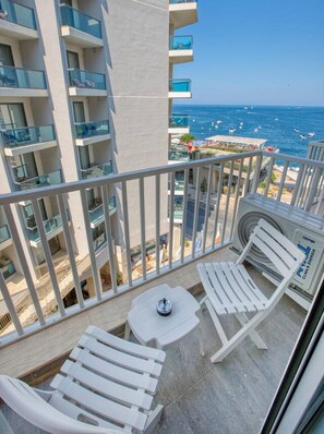 A balcony with white lounge chairs and a small table, overlooking a beach with boats and umbrellas.