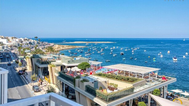 A coastal hotel with outdoor seating overlooking the sea, a road with cars, and a clear blue sky.