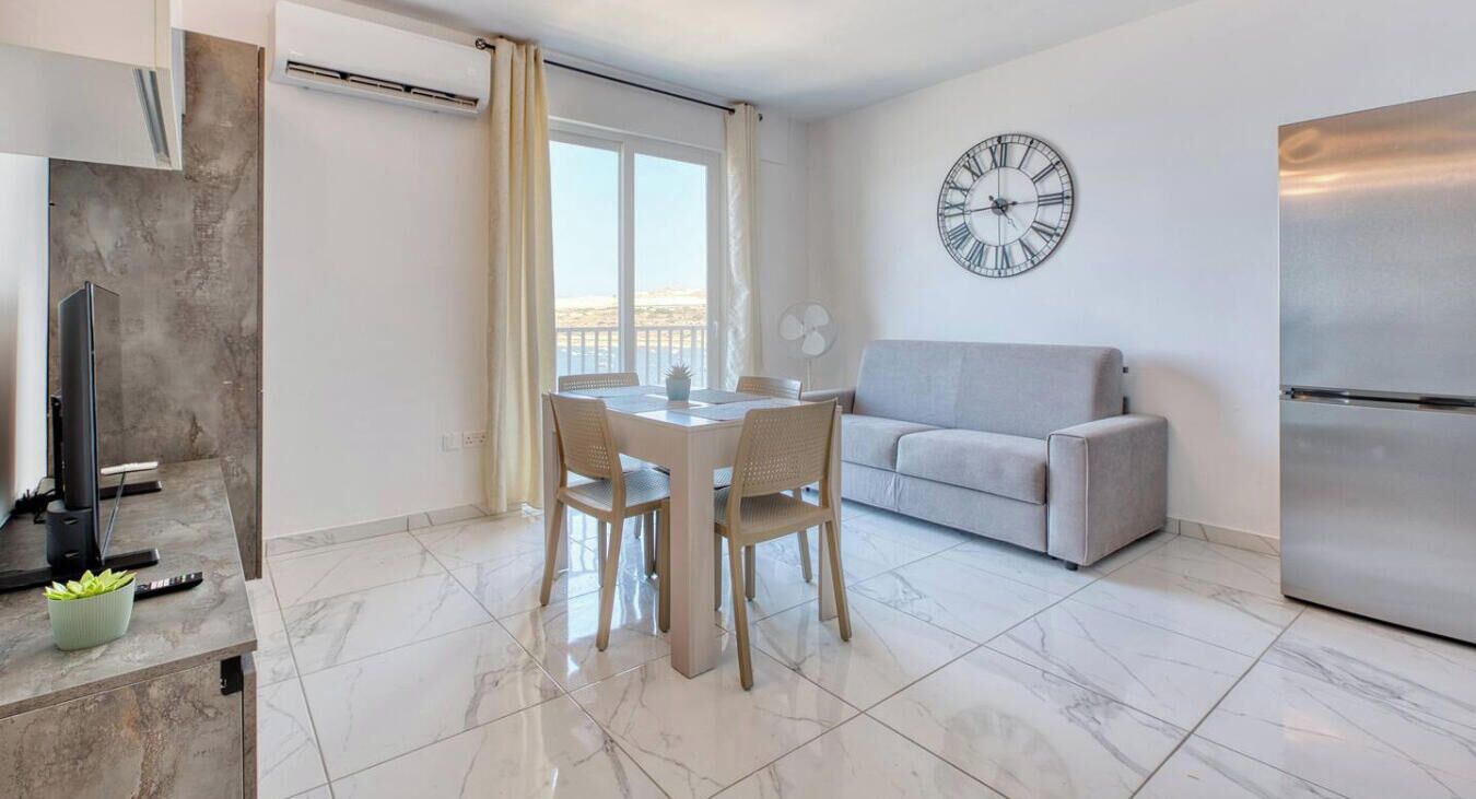 Een moderne woonkamer met een eethoek, een grijze bank, een grote wandklok en een keuken met een koelkast.