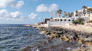 A coastal hotel with a rocky shoreline, white buildings, palm trees, and a clear sky. - Stylish 2BR Stay with Balcony Sunsets & Sea Views! (St. Paul`s Bay)