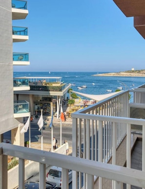 A coastal building with balconies overlooking the sea, a street with pedestrians, and a clear sky.