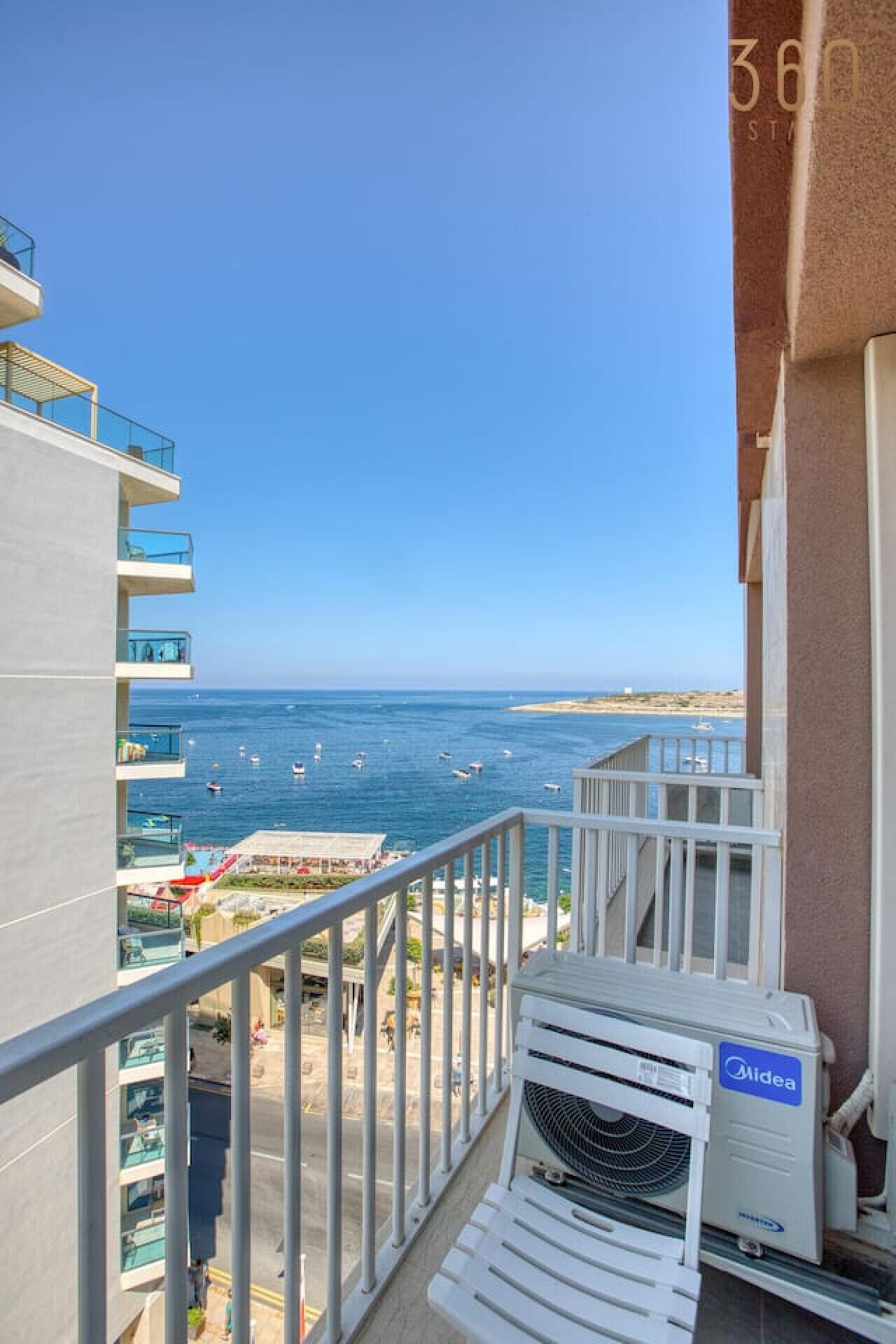A balcony with a view of the sea, a white air conditioning unit, and a clear sky.
