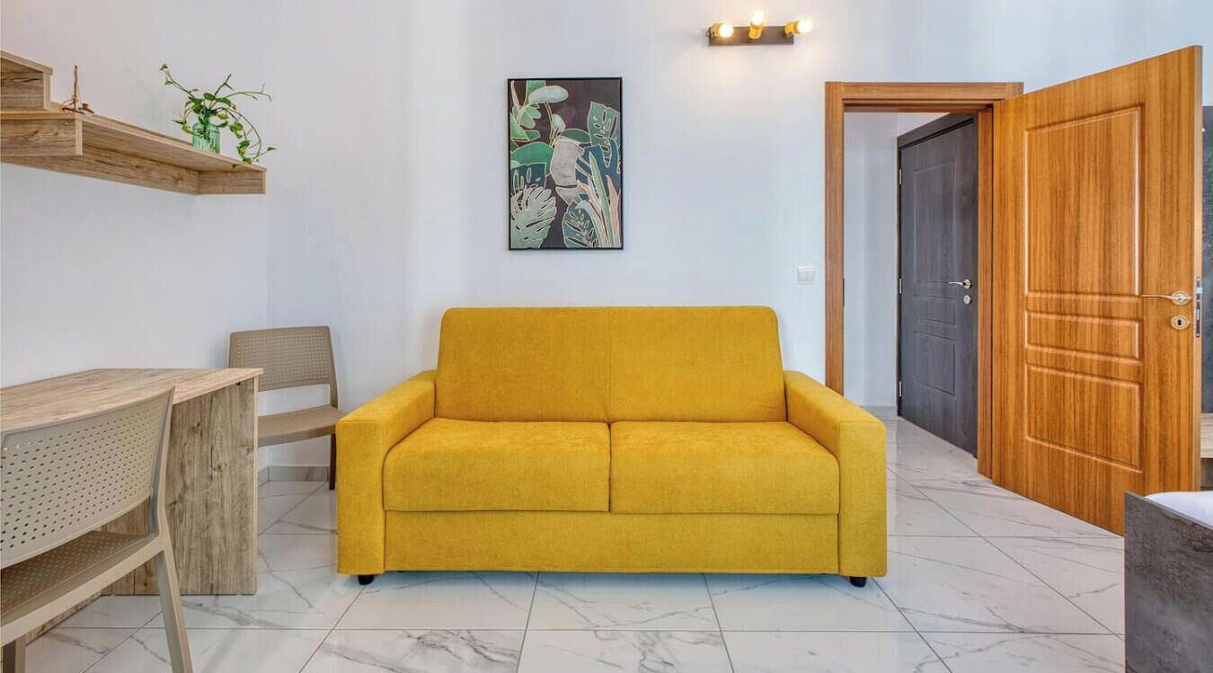 A modern living room with a yellow sofa, a wooden coffee table, and a dining area with a wooden chair. There is a painting on the wall and a door to the right.