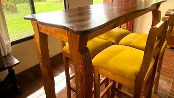Una mesa de comedor de madera con sillas con cojines amarillos, suelo de baldosas y vistas al exterior con vegetación.