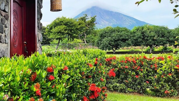 Un edificio de piedra con una puerta roja, rodeado de jardines cuidados con flores rojas, y una montaña al fondo.
