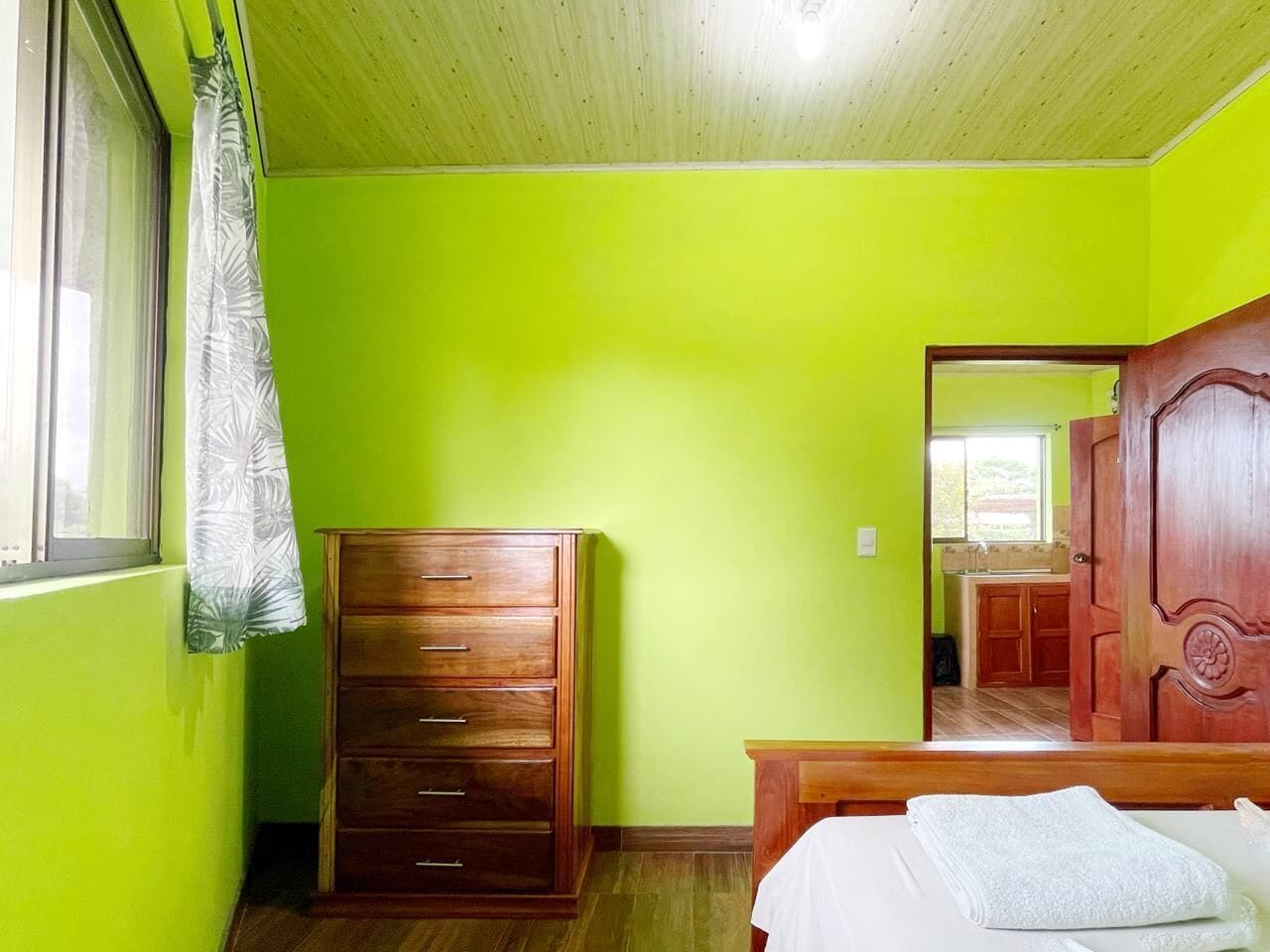 A room with a wooden dresser, a bed with a white blanket, and a wooden door leading to a kitchen.