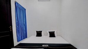 Ein kleines Zimmer mit einem Einzelbett, weißer Bettdecke, zwei schwarzen Kopfkissen und einem blauen Vorhang.
