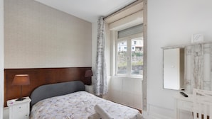 Quarto com cama, mesa de cabeceira, abajur, janela com cortinas, escrivaninha com espelho e cadeira.