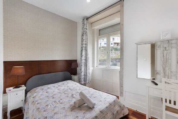 Un dormitorio con cama, mesita de noche, lámpara, ventana con cortinas, escritorio con espejo y una silla.